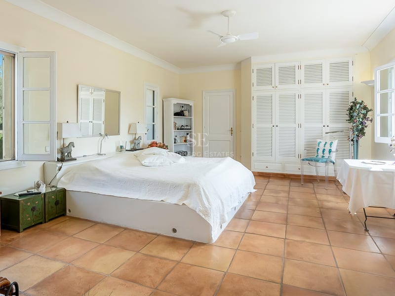 Spacious bedroom featuring terracotta floors, white louvred wardrobes, and large windows overlooking a lush garden.