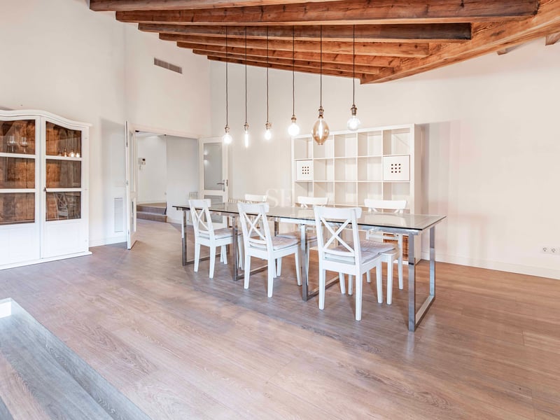 Une salle à manger spacieuse avec table en verre, chaises blanches et hauts plafonds avec poutres rustiques.