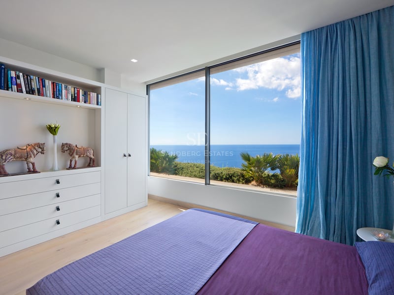 Chambre blanche moderne avec lit violet et grande baie vitrée donnant sur la mer bleue et le ciel.