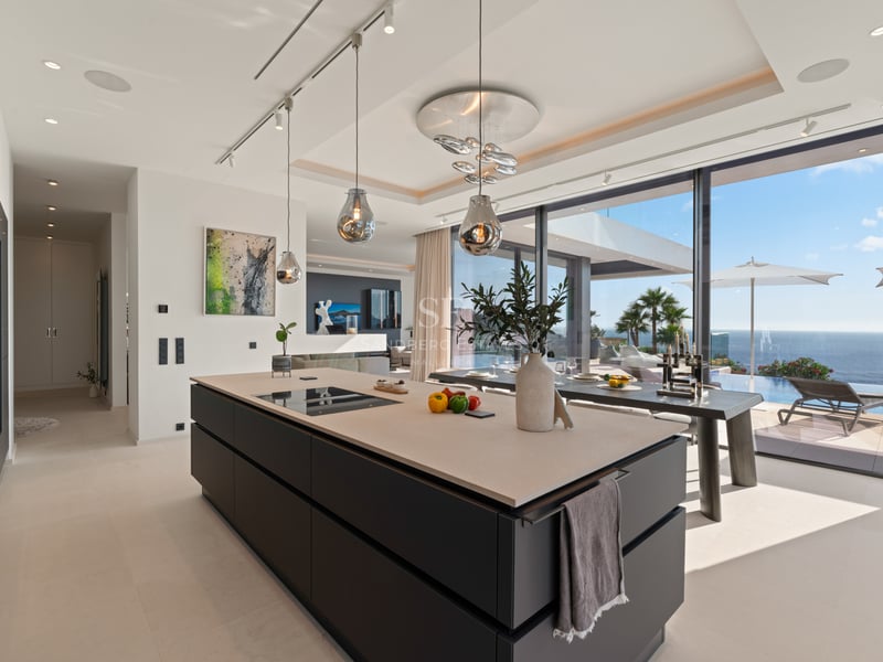 Cuisine moderne avec îlot central sombre, luminaires design en verre et baies vitrées donnant sur l'océan.