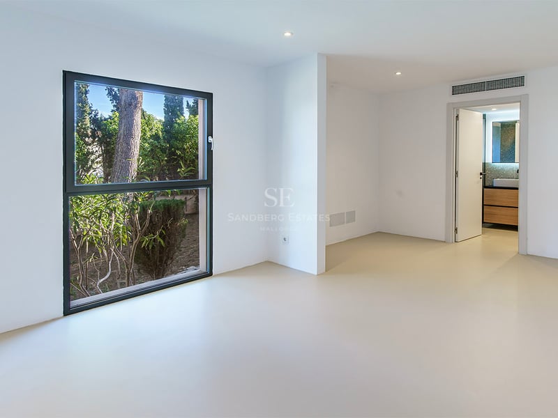 Chambre minimaliste vide avec murs blancs, sol beige, grande fenêtre sur le jardin et porte vers une salle de bain.