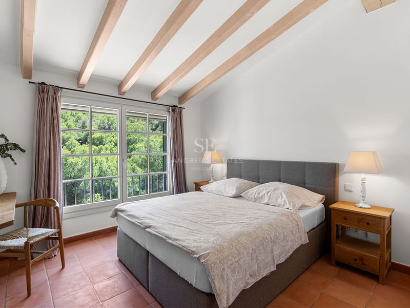 Chambre avec lit double gris, poutres apparentes, sol en terre cuite et vue sur la forêt.