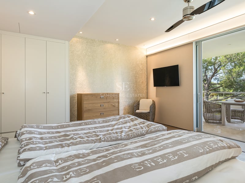 Chambre moderne avec placards blancs, commode en bois et baies vitrées ouvrant sur une terrasse ombragée par des pins.