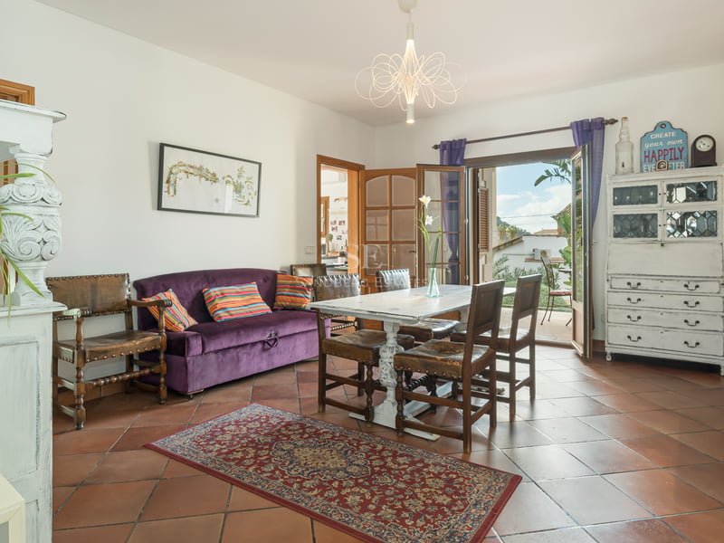 Salle à manger avec sol en terre cuite, canapé en velours violet et table en bois blanc ouvrant sur un balcon.