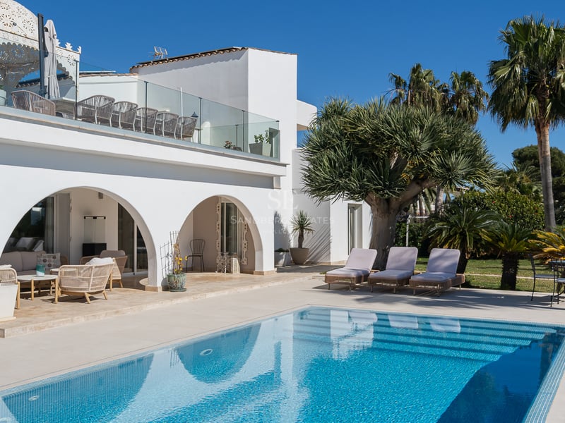 Villa blanche à arches surplombant une piscine turquoise avec chaises longues et palmiers sous un ciel bleu.
