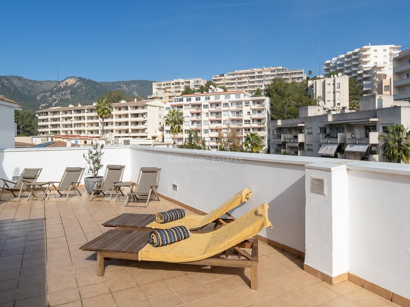 Vaste terrasse sur le toit avec chaises longues en bois, sol carrelé et vue sur les bâtiments et montagnes.