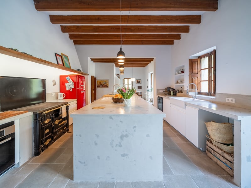 Grande cuisine avec îlot central blanc, poutres en bois apparentes, réfrigérateur rouge rétro et piano de cuisson.
