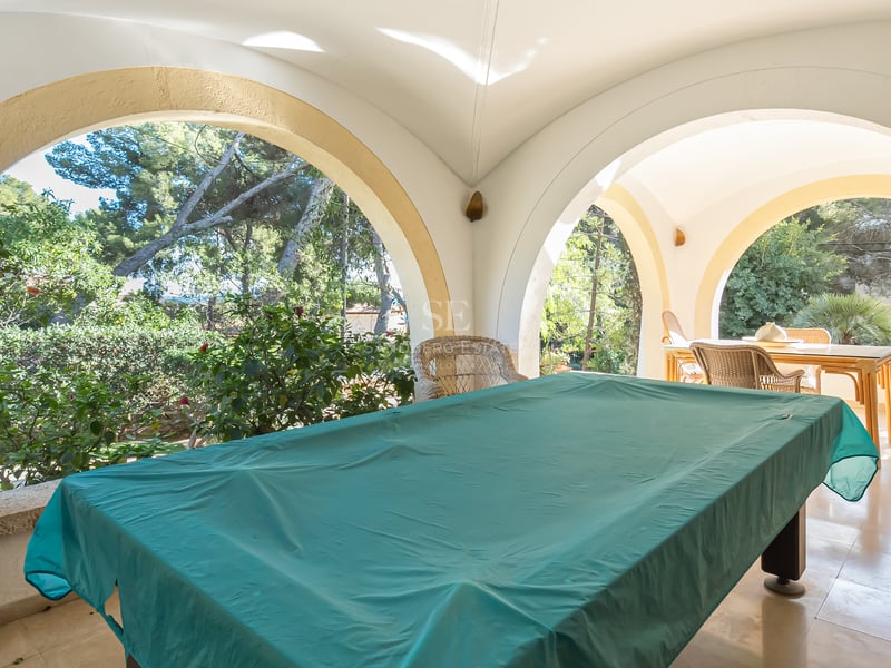 Grande terrasse couverte avec arches blanches, table de billard sous bâche verte et vue sur le jardin.