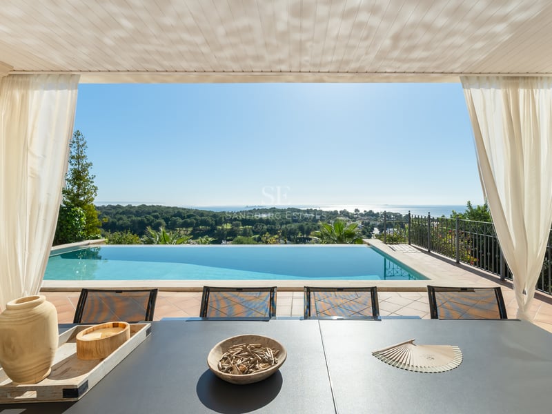 Terrasse ombragée avec table à manger surplombant une piscine à débordement, une pinède et la mer Méditerranée.