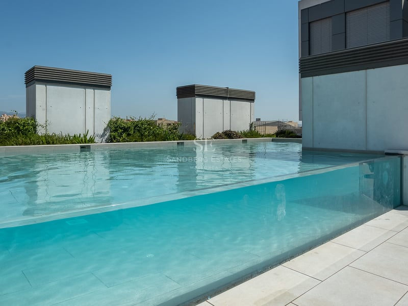 Piscine moderne sur le toit avec paroi en verre transparent et carrelage en pierre grise sous un ciel bleu.