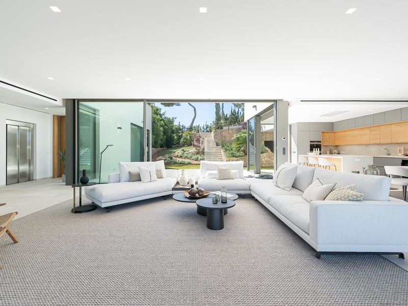 Grand salon blanc moderne avec de grands canapés, une cuisine ouverte et des baies vitrées donnant sur le jardin.