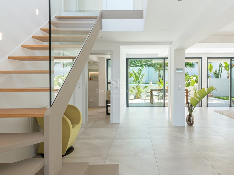 Couloir moderne avec escalier suspendu en bois et garde-corps en verre, s'ouvrant sur le salon et le jardin.