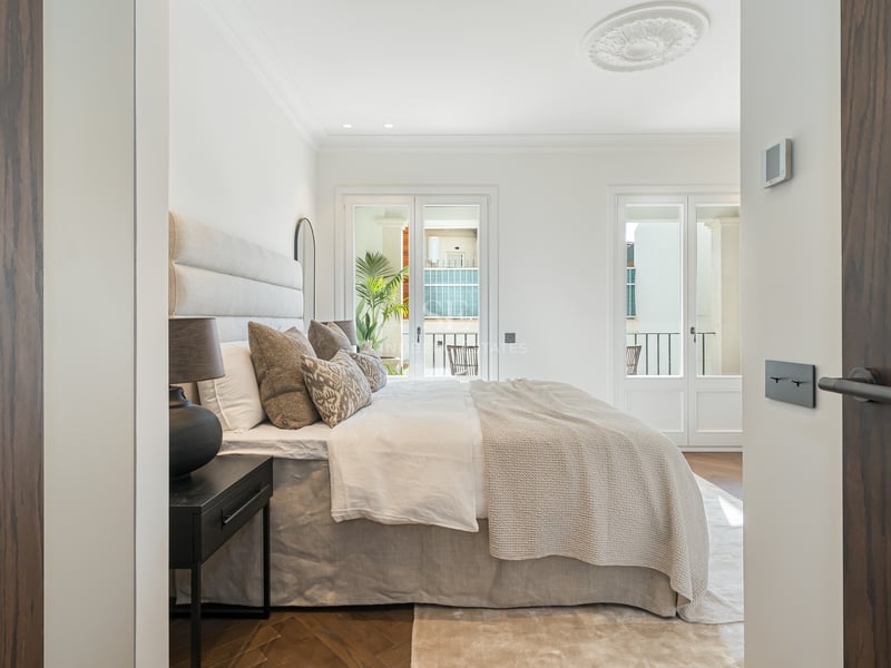Bright bedroom seen through dark wood doors, featuring a large bed, herringbone floors, and French balcony doors.