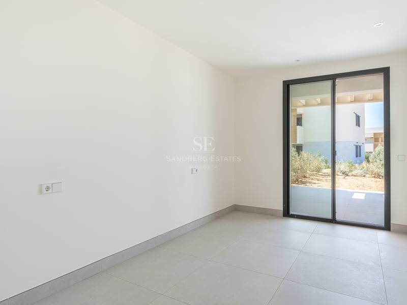 Dormitorio vacío con paredes blancas, suelo de gres gris y puerta corredera de cristal hacia un patio privado.