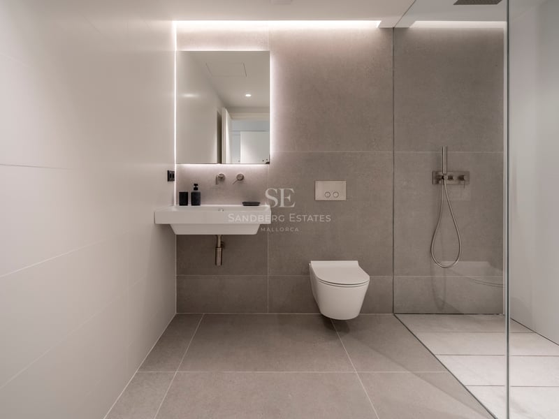 Salle de bain moderne avec carrelage gris, miroir rétroéclairé, lavabo blanc, WC suspendu et paroi de douche en verre.