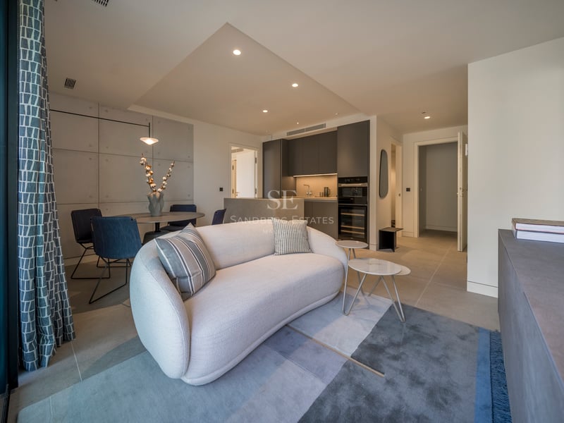 Salon moderne avec un canapé blanc incurvé, un tapis bleu et une table à manger, à côté d'une cuisine gris foncé.