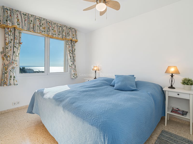 Une chambre lumineuse avec un dessus de lit bleu, vue sur la mer par une fenêtre aux rideaux fleuris et lampes de chevet.