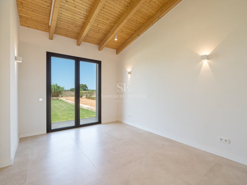 Habitación vacía con techo de vigas de madera, suelo de baldosas beige y puerta de cristal al jardín.