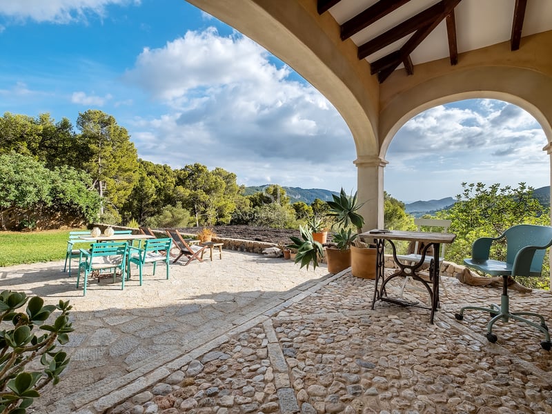 Terrasse en pierre avec arches donnant sur une vallée verdoyante et des montagnes, avec mobilier d'extérieur bleu sarcelle et bois.