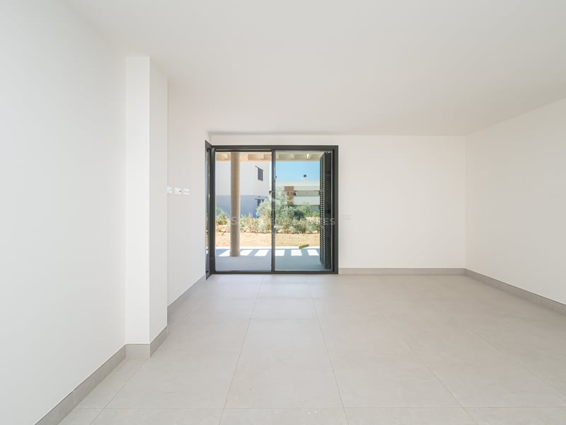 Sala vacía moderna con paredes blancas, grandes baldosas grises y puerta corredera de cristal hacia el jardín.