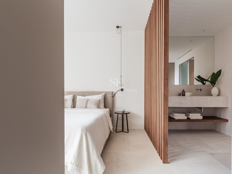 Chambre moderne avec lit beige et cloison en bois séparant un espace salle de bain en pierre.