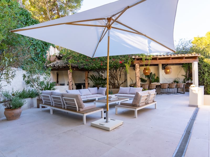 Grande terrasse avec canapés modernes, grand parasol blanc et coin repas sous une pergola entourée de verdure.