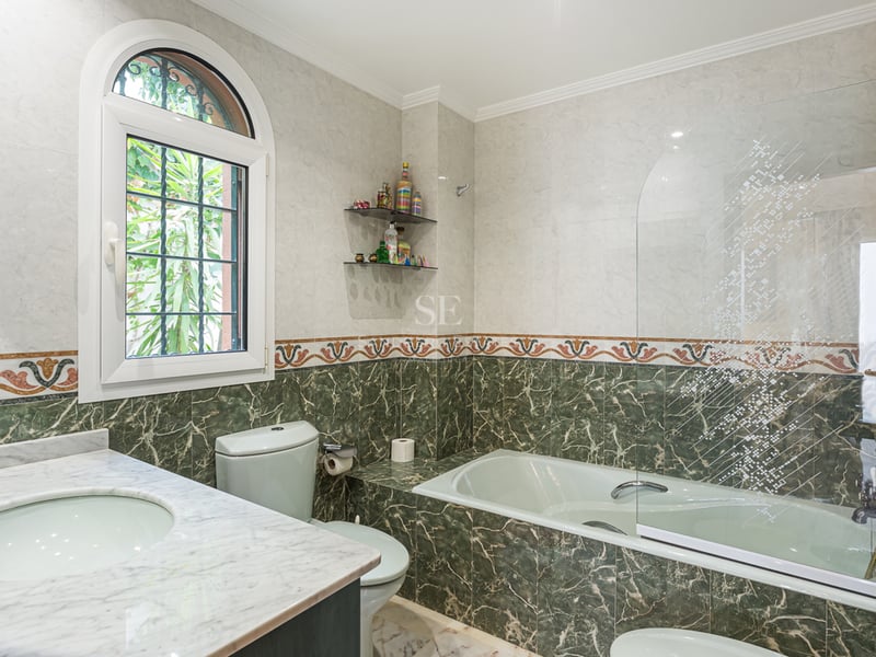 Salle de bain avec carrelage en marbre vert et blanc, baignoire avec paroi en verre et fenêtre cintrée.