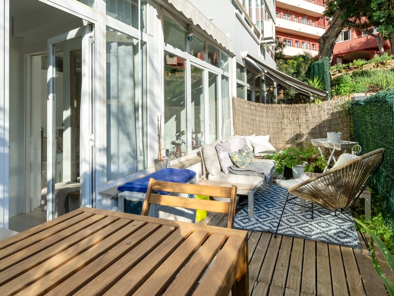 Terrasse privée en bois avec mobilier de jardin, tapis géométrique et baies vitrées sous une lumière naturelle vive.