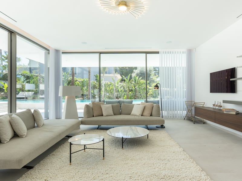 Salon contemporain avec canapés neutres, tables en marbre et grandes baies vitrées donnant sur le jardin et la piscine.