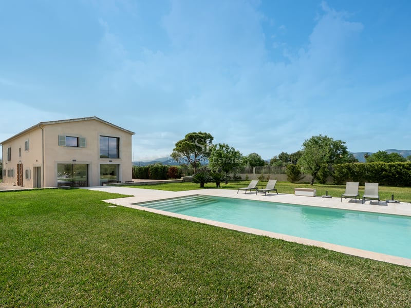 Piscine rectangulaire et chaises longues sur une pelouse verte devant une villa beige de deux étages.