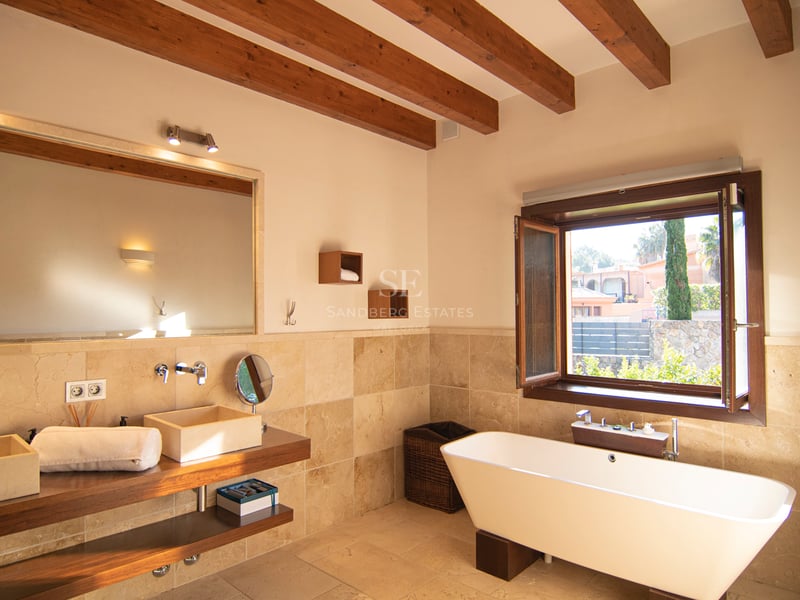 Salle de bains avec poutres apparentes, baignoire îlot, double vasque en pierre et vue sur le jardin.