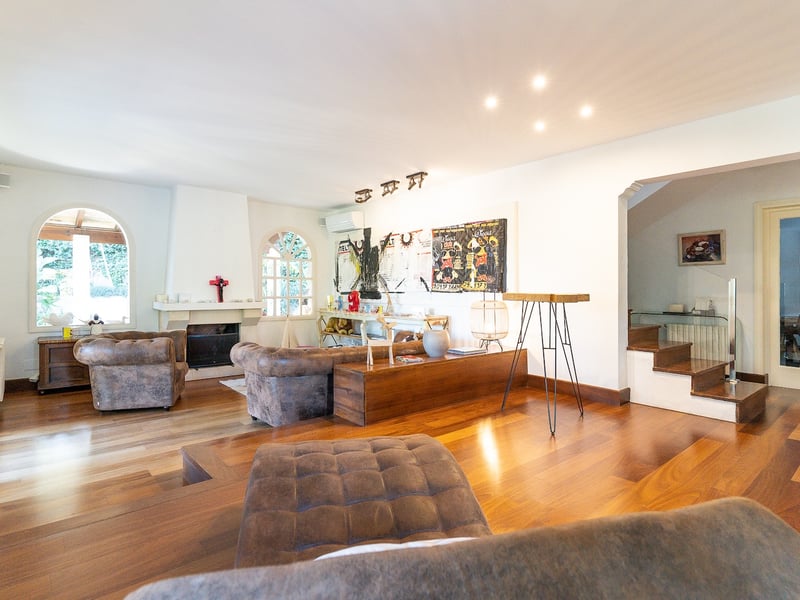 Grand salon avec parquet en bois foncé, fauteuils en cuir, cheminée et fenêtres cintrées sous un éclairage mixte.