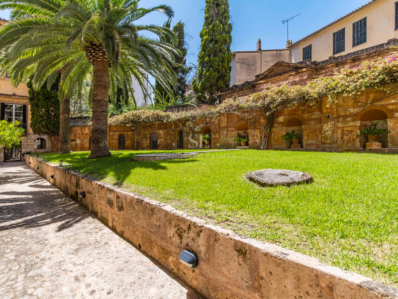 Cour ensoleillée avec pelouse, palmiers et mur en pierre classique avec niches décoratives sous un ciel bleu.