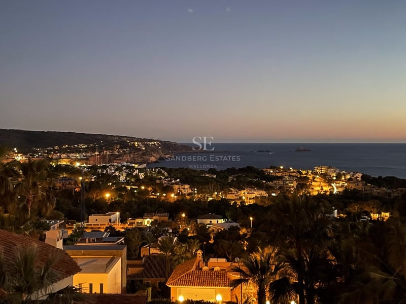 Vue nocturne surélevée d'une côte méditerranéenne avec villas éclairées, palmiers et mer sous un ciel de crépuscule.