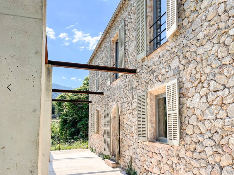 Façade en pierre naturelle d'une villa avec volets vert sauge, poutres en acier industrielles et vue sur la montagne.