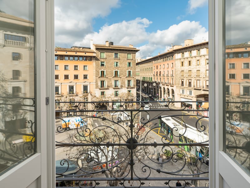 View from a classic balcony with ornate iron railing overlooking a sunlit Mediterranean city square and stone buildings.