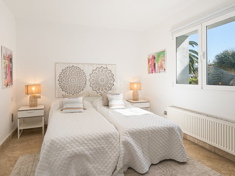 Chambre blanche avec deux lits simples, tête de lit en bois sculpté, lampes en osier et fenêtre sur le jardin.