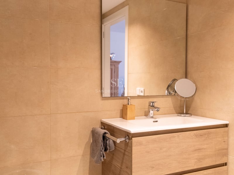 Salle de bain moderne avec meuble vasque suspendu en chêne, lavabo en céramique, grand miroir et carrelage beige.