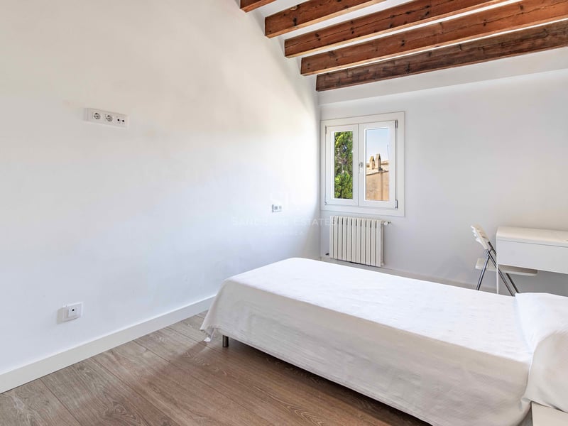 Chambre blanche avec lit simple, poutres en bois sombre au plafond incliné et parquet clair.