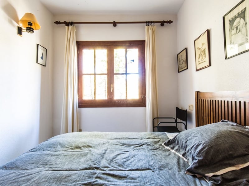 Une chambre avec un lit en bois, une couette grise et une fenêtre laissant passer une vive lumière du soleil.