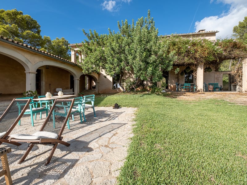 Un jardin ensoleillé avec patio en pierre, pelouse, chaises bleu sarcelle et une villa rustique aux arcades.