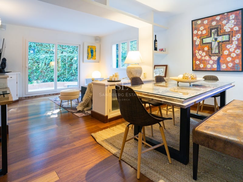 Salle à manger éclairée avec table en verre, chaises modernes et œuvre d'art, vue sur la verdure.