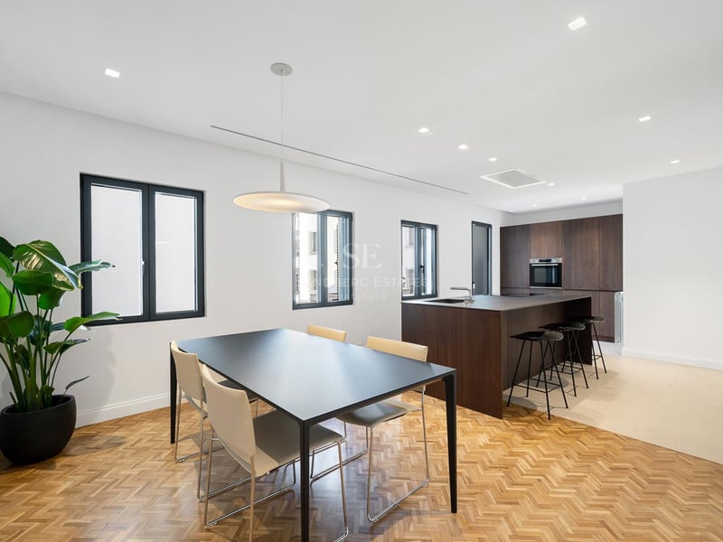 Cuisine moderne avec îlot en bois sombre et salle à manger avec parquet en chevrons et grandes fenêtres.