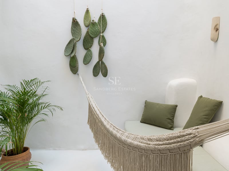 Patio aux murs blancs avec hamac à franges, coussins vert olive et plantes tropicales.