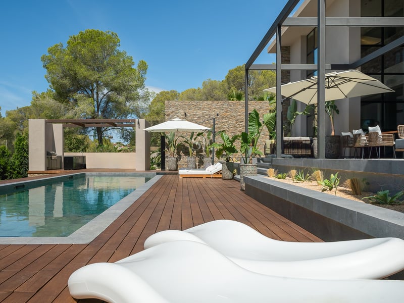 Piscine extérieure de luxe avec terrasse en bois, bains de soleil blancs et murs en pierre sous un ciel bleu.
