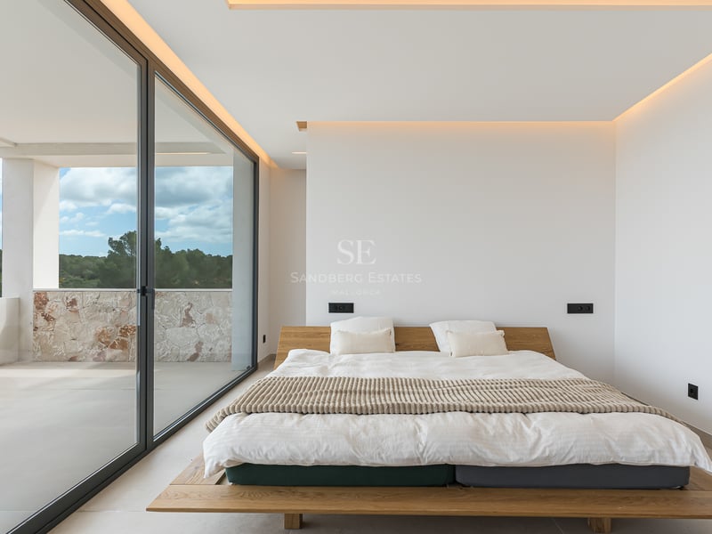 Chambre principale minimaliste avec lit en bois, baies vitrées et accès à un balcon en pierre naturelle.