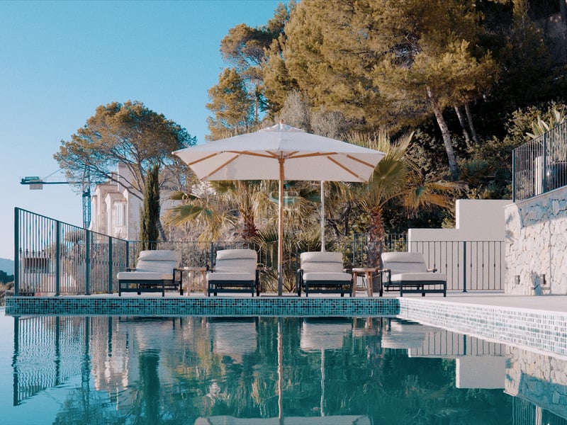 Piscine à débordement avec quatre chaises longues sous un parasol blanc, entourée de murs en pierre et de pins.