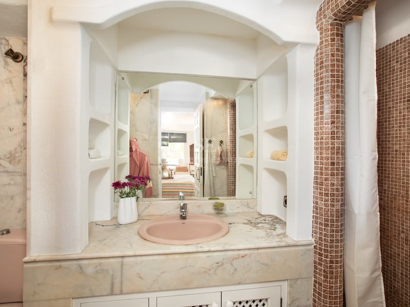 Salle de bain avec vasque rose, plan en marbre, miroir arqué et colonne en mosaïque brune.