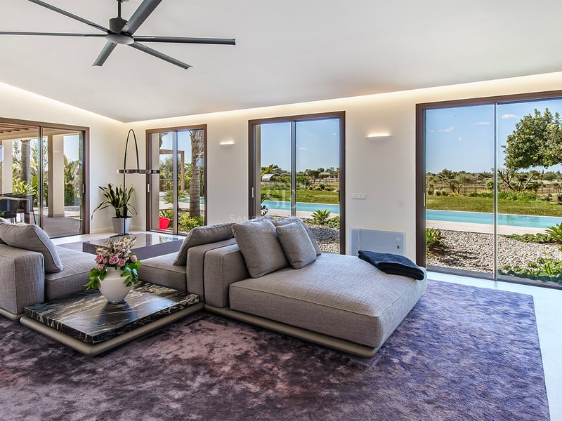 Salon moderne avec canapé modulaire gris, tables en marbre et grandes baies vitrées donnant sur un jardin avec piscine.