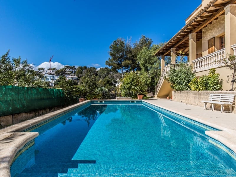 Grande piscine rectangulaire aux eaux turquoises à côté d'une villa méditerranéenne avec terrasse ensoleillée et ciel bleu.
